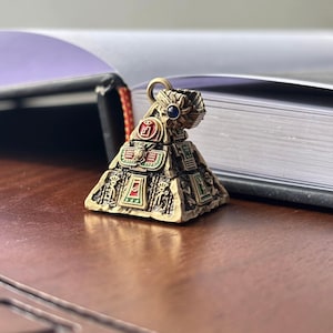 Può includere: Ciondolo a forma di piramide in stile antico, color oro, con dettagli in smalto rosso, verde e blu. Presenta intagli intricati e un piccolo anello per l'attacco. La piramide misura circa 2,5 cm di altezza.