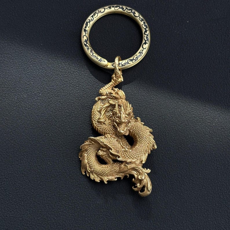 May include: A gold-colored metal keychain featuring a detailed dragon pendant. The dragon is intricately designed with scales, claws, and a flowing tail. The keychain ring has a decorative pattern.
