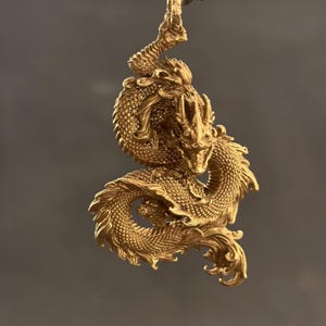 May include: A detailed, golden dragon pendant with intricate scales, horns, and a flowing mane. The dragon is suspended against a neutral background, showcasing its three-dimensional design. The pendant's metallic finish reflects light, highlighting its ornate features.