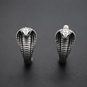 May include: A pair of silver-toned metal earrings shaped like cobra snakes. Each earring features a detailed, textured scale pattern on the hood and head of the cobra, with a dark, oxidised finish that highlights the intricate snake design.