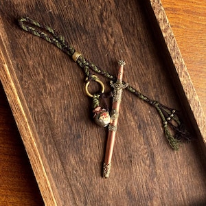 Op de afbeelding: Een miniatuur zwaardhanger met een koperkleurig lemmet en een sierlijke goudkleurige greep. De hanger is bevestigd aan een gevlochten koord in groen- en bruintinten, met een kleine decoratieve bedel en een goudkleurige ring.