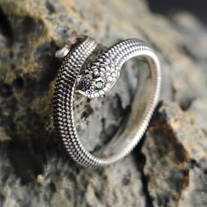 Può includere: Un anello color argento a forma di serpente avvolto con una superficie testurizzata simile a scaglie. La testa del serpente presenta una piccola pietra preziosa rotonda di colore verde come occhio. Il metallo ha una finitura ossidata che risalta.