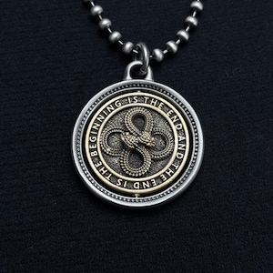 May include: A silver-toned pendant necklace with a circular pendant. The pendant features a detailed design of two intertwined snakes in a bronze color, surrounded by the words "THE BEGINNING IS THE END AND THE END IS THE BEGINNING".