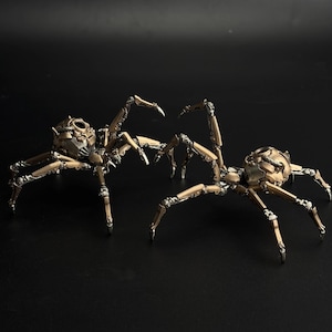May include: Two steampunk-style spider sculptures. The spiders are bronze and silver in color, with intricate mechanical details. Each spider has a rounded head and multiple articulated legs. The sculptures are set against a black background.