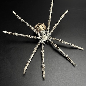 May include: A detailed, metallic spider sculpture with articulated legs and a segmented body. The spider features a gold-colored head and a silver-toned body, set against a dark background. The intricate design suggests a steampunk or fantasy theme.