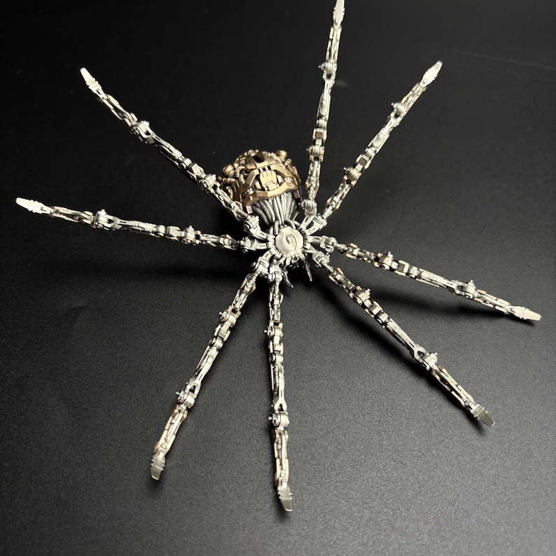 May include: A detailed, metallic spider sculpture with articulated legs and a segmented body. The spider features a gold-colored head and a silver-toned body, set against a dark background. The intricate design suggests a steampunk or fantasy theme.