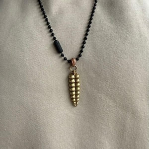 May include: A necklace featuring a matte black beaded chain with a cylindrical black bead accent and a metallic gold-toned pendant shaped like a segmented rattlesnake tail. The pendant is attached to the chain by a small copper-colored jump ring.