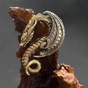 Può includere: Una dettagliata statuetta di drago bicolore. Il drago è posato su una superficie marrone ruvida. La testa e il corpo del drago sono dorati, mentre le ali sono argentate. La coda del drago si arriccia a spirale.