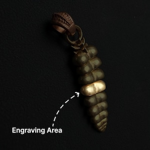 May include: A dark brown, segmented rattlesnake rattle pendant featuring a single smooth, metallic gold-colored band in the center. A dashed white arrow points to this gold band, which is labeled with the text Engraving Area below it.