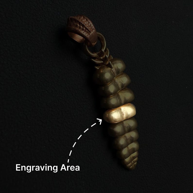 May include: A dark brown, segmented rattlesnake rattle pendant featuring a single smooth, metallic gold-colored band in the center. A dashed white arrow points to this gold band, which is labeled with the text Engraving Area below it.
