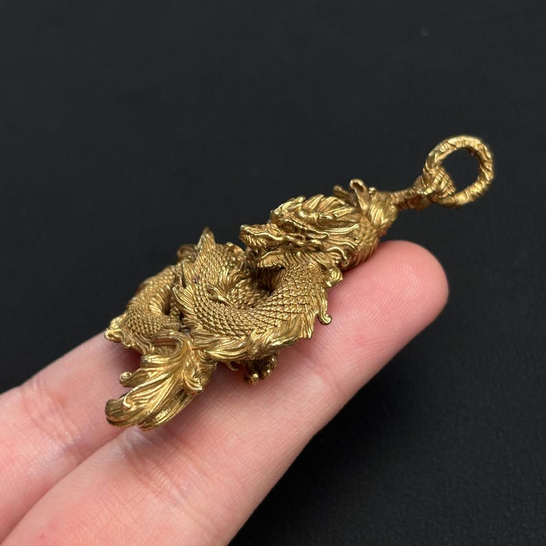 May include: A detailed brass dragon pendant with intricate scales and a looped top for hanging. The dragon is in a coiled pose, showcasing its detailed features. The pendant is a warm gold color.