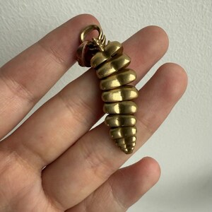 May include: A gold-toned metal pendant shaped like a rattlesnake rattle, featuring a series of segmented, rounded, and tapered horizontal ridges that decrease in size toward the pointed tip, with a small circular loop at the top for attachment.
