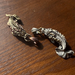 Könnte beinhalten: Zwei detaillierte Koi-Fisch-Anhänger. Einer ist goldfarben mit rosa Akzenten, der andere silberfarben mit goldenen Akzenten. Beide haben aufwendige Schuppen- und Flossendetails. Die Anhänger liegen auf einer Holzoberfläche.