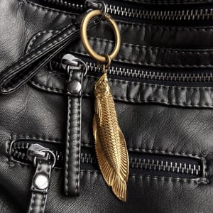 Op de afbeelding: Een goudkleurige visbedel hangt aan een messing ring, bevestigd aan een zwarte leren tas. De visbedel heeft gedetailleerde schubben en vinnen. De tas heeft meerdere ritsen en stikseldetails.