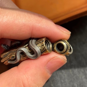May include: A detailed, silver snake coils around a brass log-shaped container. The container has a removable cap with a black interior and gold trim. The snake's head is visible, adding to the intricate design.
