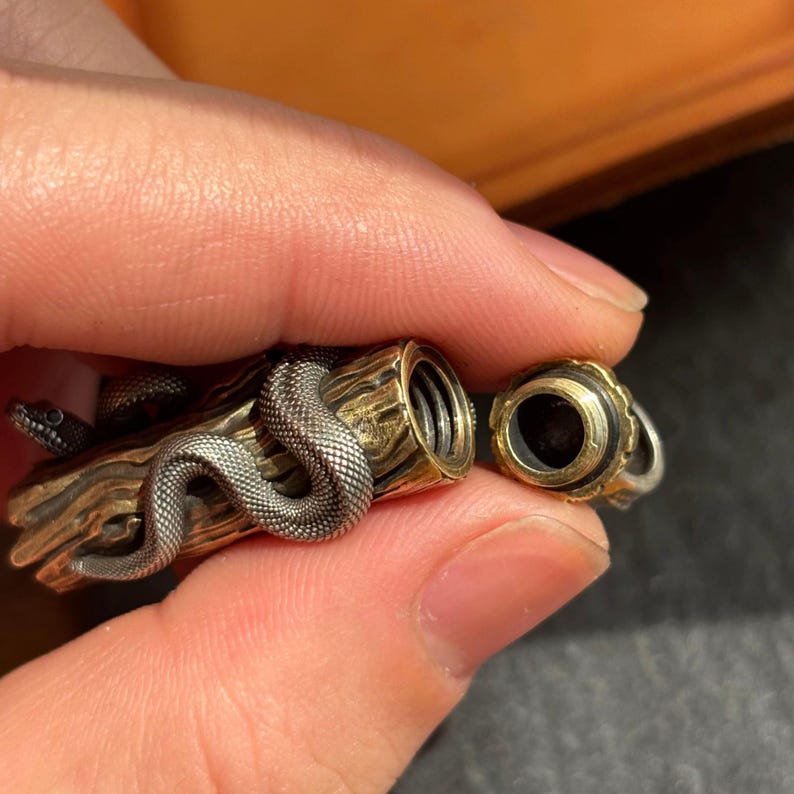 May include: A detailed, silver snake coils around a brass log-shaped container. The container has a removable cap with a black interior and gold trim. The snake's head is visible, adding to the intricate design.