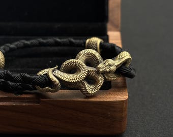 May include: A black braided bracelet with a detailed gold-colored snake clasp. The bracelet is displayed inside a wooden box with a black velvet interior. The snake clasp features intricate scales and coiled design.