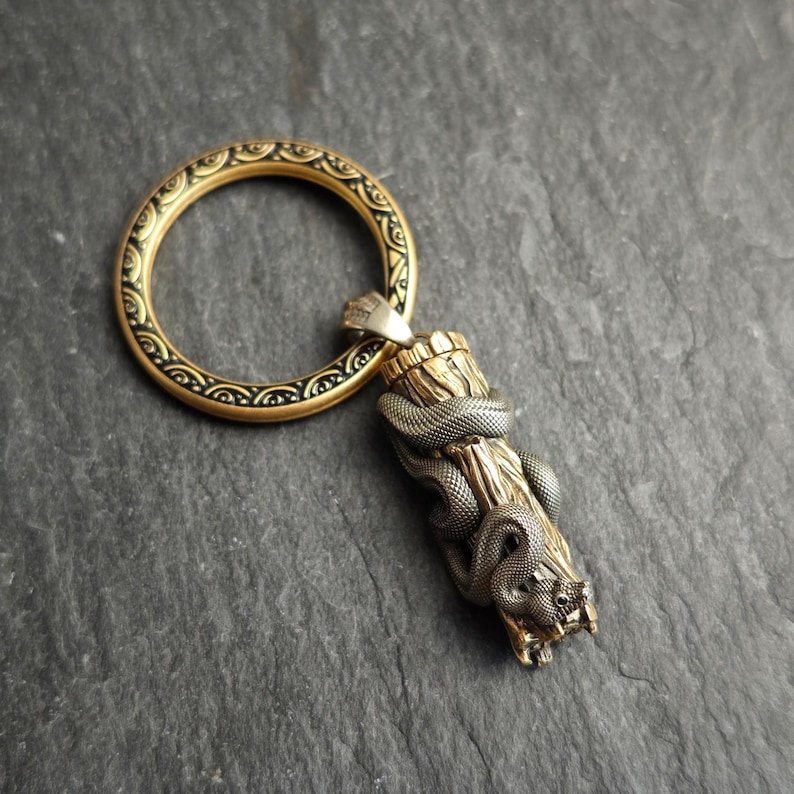 May include: A bronze keychain with a snake design wrapped around a wooden post. The keychain has a decorative gold ring with black detailing. The snake is silver, and the post is light brown. The overall design is intricate.
