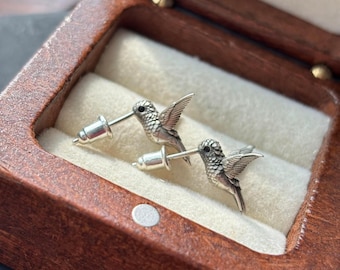May include: A pair of silver hummingbird stud earrings displayed in a wooden jewelry box. The earrings feature detailed bird designs with outstretched wings and a post and backing. The box has a dark brown finish and a cream-colored interior.