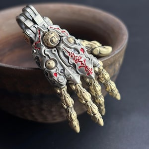 May include: A detailed, metallic hand pendant with intricate mechanical and artistic elements. The pendant features a silver-colored base with gold-colored accents and red detailing. The design incorporates a blend of industrial and artistic styles, with visible joints and engraved patterns.