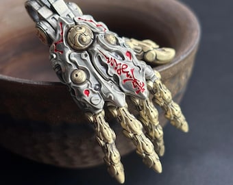 May include: A detailed, metallic hand pendant with intricate mechanical and artistic elements. The pendant features a silver-colored base with gold-colored accents and red detailing. The design incorporates a blend of industrial and artistic styles, with visible joints and engraved patterns.