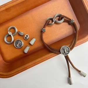 Op de afbeelding: Een bruine bolo stropdas met een zilveren levensboomhanger en decoratieve zilveren accenten. De afbeelding toont ook diverse zilveren kralen en een slangvormige bedel, allemaal gepresenteerd in een bruine bak.
