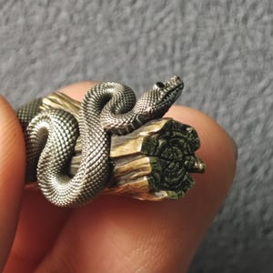 May include: A detailed metal sculpture of a snake coiled around a piece of wood. The snake is silver-coloured with textured scales, and the wood has a natural, weathered appearance. The sculpture is held in a hand.