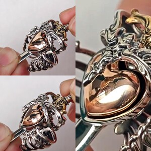 Op de afbeelding: Een metalen haaraccessoire met een gepolijst ros&eacute;gouden hart in het midden, ingesloten in een gedetailleerde zilverkleurige kooistructuur die lijkt op een borstkas. Het ontwerp bevat ingewikkelde, vloeiende metaalbewerking en een gladde afwerking.