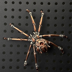 May include: A detailed, steampunk-style metal spider sculpture. The spider features a copper-colored body and legs with intricate mechanical details, including rivets and articulated joints. The legs are a mix of copper and silver, and the spider is set against a black background.