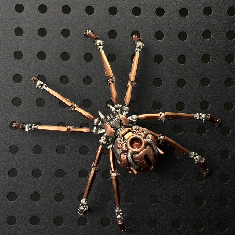 May include: A detailed, steampunk-style metal spider sculpture. The spider features a copper-colored body and legs with intricate mechanical details, including rivets and articulated joints. The legs are a mix of copper and silver, and the spider is set against a black background.