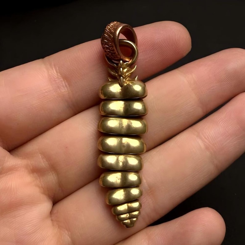 May include: A gold-toned pendant shaped like a rattlesnake rattle, featuring a series of segmented, rounded horizontal ridges that taper toward the bottom, topped with a textured, circular bail for attachment to a chain or cord.