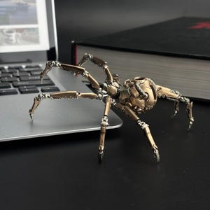 May include: A detailed, bronze-colored mechanical spider sculpture with articulated legs and body segments. The sculpture is posed on a laptop, with a book in the background. The spider's intricate design suggests a steampunk or industrial aesthetic.