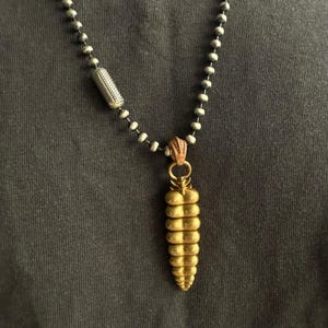 May include: A necklace featuring a metallic ball chain with a cylindrical textured bead and a gold-toned pendant shaped like a segmented rattlesnake tail. The pendant hangs from a decorative bail, and the chain consists of small, uniform spheres.