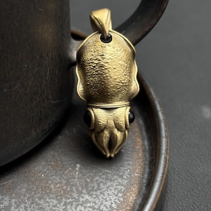 Peut inclure: Pendentif en laiton représentant un calmar, avec des détails texturés et des yeux noirs. Le pendentif est suspendu à une boucle, présentant un design unique sur le thème marin.