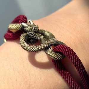May include: A bracelet featuring a metallic gold-toned snake charm with detailed scales and small horns. The snake is coiled around two strands of deep burgundy cord, which are woven into a textured, rope-like design that secures the charm.