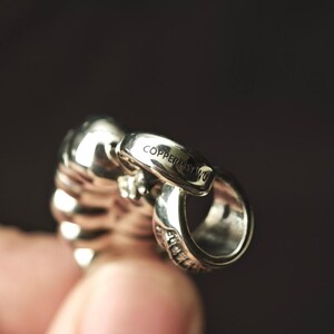 May include: A polished silver-toned metal pendant featuring a curved, ribbed design with a prominent bail. The word COPPERLISTANU is clearly engraved on the top surface of the bail, which connects to a rounded, textured lower section of the piece.