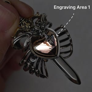 Op de afbeelding: Een zilverkleurige metalen hanger met een ingewikkeld, sierlijk frame rondom een centrale hartvormige, lichtbruine edelsteen. Het ontwerp bevat een puntige, zwaardachtige verlenging aan de onderkant. De tekst luidt Engraving Area 1.