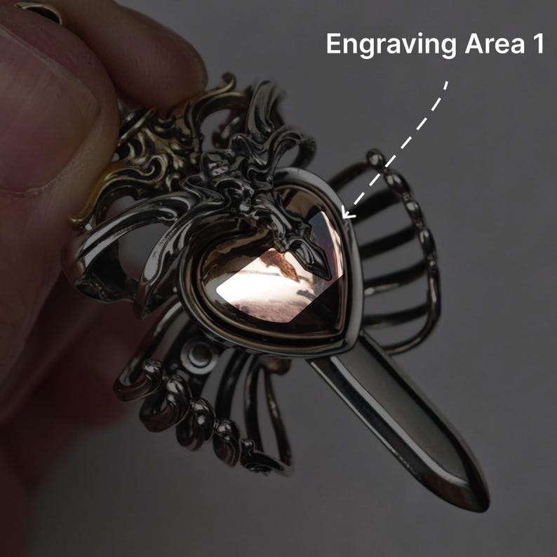 Op de afbeelding: Een zilverkleurige metalen hanger met een ingewikkeld, sierlijk frame rondom een centrale hartvormige, lichtbruine edelsteen. Het ontwerp bevat een puntige, zwaardachtige verlenging aan de onderkant. De tekst luidt Engraving Area 1.