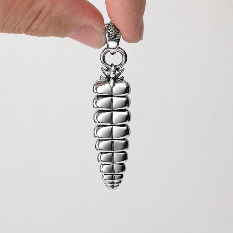 May include: A silver-toned metal pendant shaped like a rattlesnake tail rattle. The pendant features a series of segmented, rounded horizontal layers that taper toward the bottom, topped with a textured bail for attachment to a chain or cord.