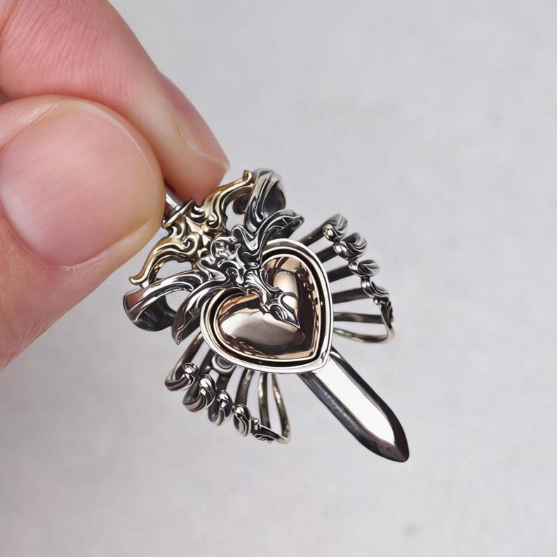 Op de afbeelding: Een metalen hanger met een centraal gepolijst hart in een warme ros&eacute;gouden tint, omringd door ingewikkeld zilverkleurig filigraan metaalwerk dat een zwaardachtige structuur vormt die verticaal achter het hartelement uitsteekt.