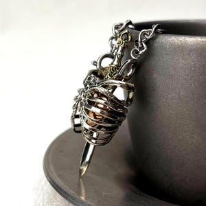 Op de afbeelding: Een zilverkleurige metalen hanger met een skeletachtige ribbenkaststructuur die een gladde, goudkleurige metalen bol omsluit. De hanger is bevestigd aan een zilveren ketting en heeft een dolkvormig element aan de onderkant van de kooi.