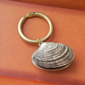 Puede incluir: Un llavero de metal en tono latón con una anilla circular y un dije de concha de almeja plateada con textura. La concha presenta crestas concéntricas detalladas y un aspecto realista y desgastado. La anilla de metal es lisa y circular.