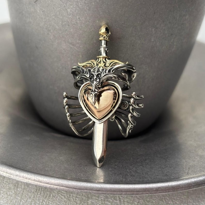 Op de afbeelding: Een zilver- en goudkleurige metalen hanger met een centraal hart dat wordt doorboord door een verticaal zwaard. Het ontwerp bevat ingewikkelde ribachtige structuren aan de zijkanten, met een kleine schedel aan de bovenkant van het gevest.