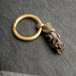 May include: A gold-coloured key ring with a detailed bronze snake wrapped around a wooden log. The key ring is made of brass. The snake is a dark, metallic bronze. The log has a textured, natural appearance.