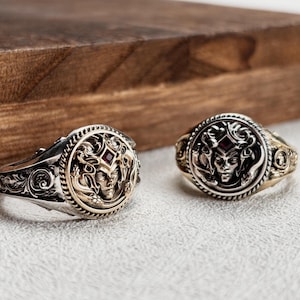 Puede incluir: Dos anillos ornamentados con un diseño de rostro tipo Medusa. Los anillos presentan una banda plateada con detalles dorados alrededor de la cara y una pequeña gema roja. Los anillos están sobre una superficie clara con un fondo de madera.