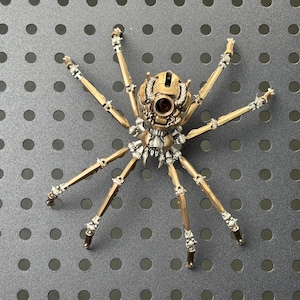 May include: A detailed, mechanical spider sculpture with a bronze and silver finish. The spider has eight articulated legs and a complex body design, featuring intricate gears and rivets. The sculpture is set against a gray background.