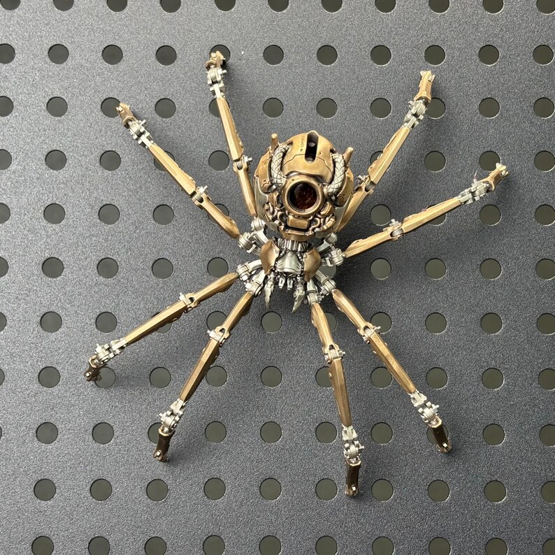 May include: A detailed, mechanical spider sculpture with a bronze and silver finish. The spider has eight articulated legs and a complex body design, featuring intricate gears and rivets. The sculpture is set against a gray background.