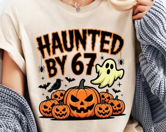 May include: Beige t-shirt with a Halloween-themed graphic. The design features the text "HAUNTED BY 67" in orange and black, along with jack-o'-lanterns, a ghost, bats, and spiders. The shirt is a casual, short-sleeved style.