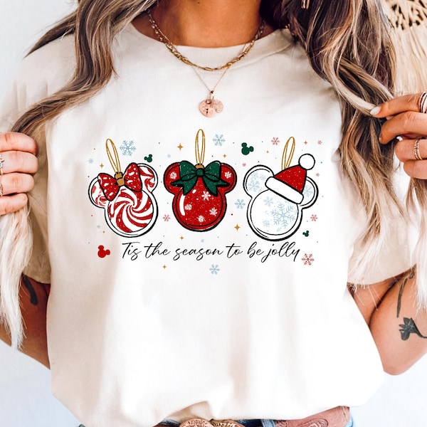 Könnte beinhalten: Cremefarbenes T-Shirt mit drei Mickey Mouse-förmigen Weihnachtsschmuckstücken. Die Ornamente sind rot, weiß und grün, mit Schneeflocken- und Zuckerstangendetails. Der Text "Tis the season to be jolly" ist darunter gedruckt.