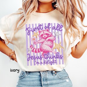 May include: Ivory-colored t-shirt featuring a pink raccoon illustration holding a flower, with text reading "First of all Jesus thinks I'm a delight." The design includes purple stripes and gold stars.